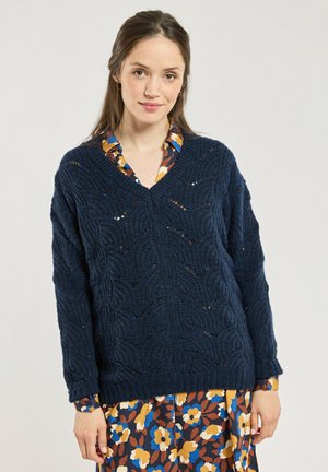 Jeune femme aux longs cheveux portant un pull en maille bleu marine superposé sur une robe fleurie avec des fleurs jaunes, bleues et marron.