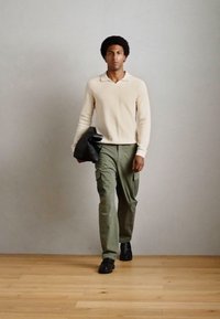Beige svetr, olivové cargo kalhoty a černé boty. Svetr má lehkou texturu látky a jemný límec. Model drží černou bundu.