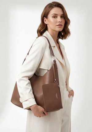Vrouw in witte jas houdt een grote bruine leren tote bag met bijpassend aangehecht tasje vast, staand tegen een effen witte achtergrond.