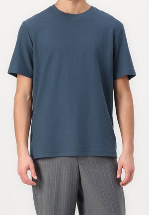 Homme portant un t-shirt bleu uni à manches courtes et col rond, et un pantalon gris à fines rayures, debout devant un fond blanc.
