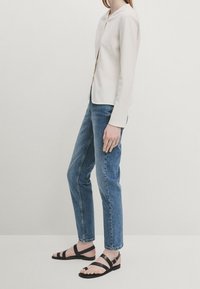 Light beige fitted jacket, blue denim jeans, and black strappy sandals. The jacket features a rounded neckline and textured fabric.