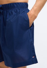 Άνδρας που φοράει μπλε σορτς με ελαστική μέση, με το χέρι στην δεξιά τσέπη, μπροστά σε μονόχρωμο λευκό φόντο.