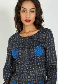 Mujer con cabello rizado y oscuro y lápiz labial rojo, vistiendo un vestido estampado con bordado floral azul y mangas largas.
