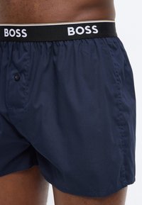 Σκούρα μπλε ανδρικά εσώρουχα μποξέρ με μαύρη ελαστική μέση με λευκά λογότυπα "BOSS", φορεμένα από άτομο με σκούρο τόνο δέρματος.