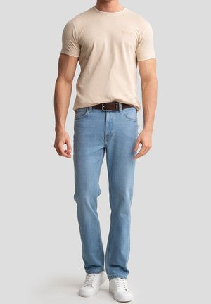 Mand iført beige t-shirt, lyseblå jeans med brun bælte og hvide sneakers, stående mod en ensfarvet lys grå baggrund.