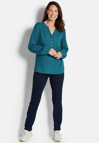 Blusa teal con scollo a V e lista di bottoni, abbinata a jeans scuri e scarpe da ginnastica bianche. Tessuto liscio, maniche lunghe e polsini arrotolati.