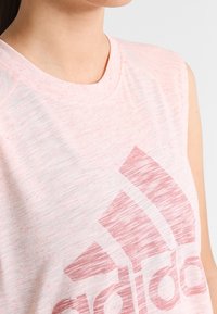 Débardeur de sport rose clair en tissu doux et texturé, avec un grand logo rouge estompé sur le devant.