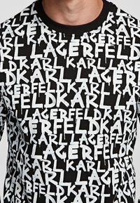 Svart sweatshirt täckt av vit text som upprepas med "KARL LAGERFELD," med rund hals och ribbade muddar, tillverkad av ett mjukt material.