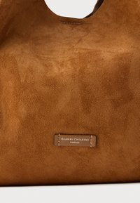 Hnědá semišová kabelka s hladkou texturou, vybavená výraznou značkovou visačkou připevněnou na boku. Zaobřený horní otvor a minimalistický design.