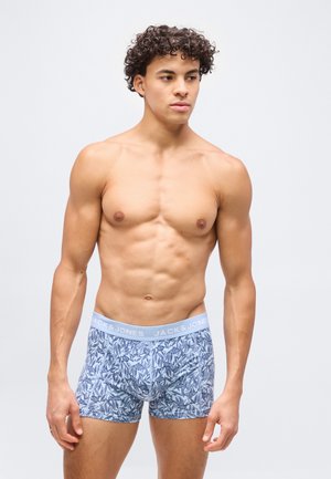 Bărbat tânăr și musculos cu păr creț, purtând boxeri albaștri cu model și talie "JACK & JONES", stând pe un fundal simplu.