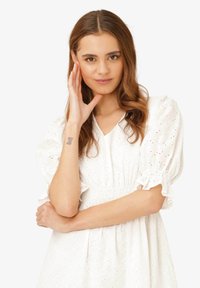 Weißes Augenlederkleid mit geraffter Taille, kurzen Puffärmeln und Rüschensäumen. Das Modell hat lange, wellige Haare und ein Tattoo auf ihrem Arm.