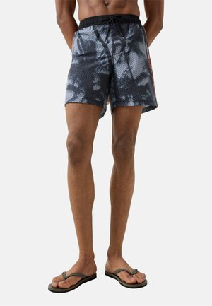 Mand iført sorte og grå tie-dye badebukser med sort linning og sorte klip-klappere, stående foran en ensfarvet baggrund.