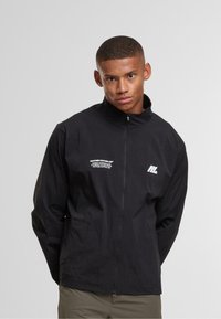 Giovane uomo che indossa una giacca nera con zip e testo bianco del logo sul lato sinistro del petto, in piedi contro uno sfondo grigio uniforme.