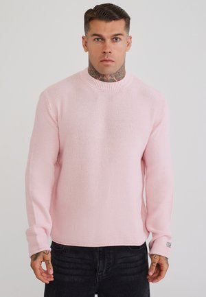 Mand med hals- og håndtatoveringer iført en lyserød tekstureret sweater og mørke jeans, stående mod en ensfarvet lys baggrund.
