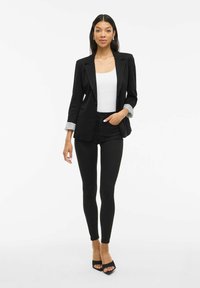 Frau in schwarzem Blazer mit hochgerollten gestreiften Ärmeln, weißem Tanktop, schwarzen Skinny-Hosen und schwarzen offenen High Heels, die vor einem weißen Hintergrund steht.