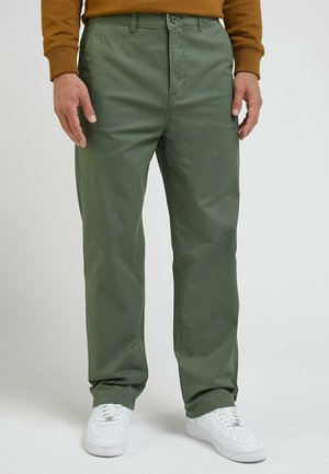 Homme portant un pantalon droit vert olive, des baskets blanches et une chemise marron à manches longues, debout devant un fond uni.