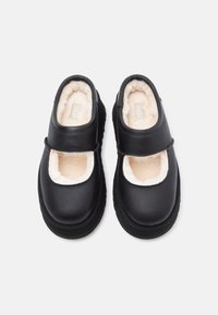 Sapatos pretos de couro sem atacadores com solas grossas e forro de pelúcia em tom creme, apresentando uma tira larga na parte superior para um ajuste seguro.