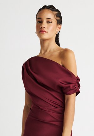 Γυναίκα με πλεξούδες στα μαλλιά που φοράει σκούρο κόκκινο σατέν φόρεμα με έναν ώμο και ρυτιδωτό κοντό μανίκι, σε απλό φόντο.