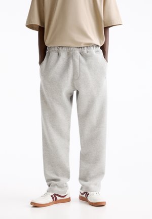 Personne portant un pantalon de survêtement gris clair avec une taille élastique et des baskets blanches à semelles marron, se tenant les mains dans les poches.