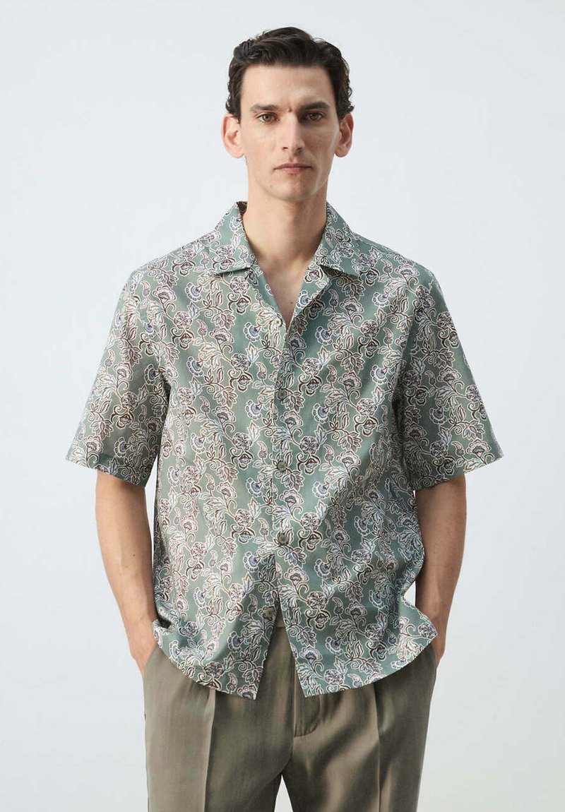 Homme portant une chemise verte à manches courtes boutonnée avec un motif floral blanc et marron et un pantalon vert olive, debout les mains dans les poches.
