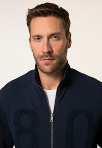 Chaqueta con cremallera en azul marino que presenta un cuello alto, ribete interior blanco en contraste y un diseño texturizado del número "80" en la parte delantera. Material suave con un acabado liso.