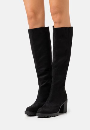 Bottes noires montantes jusqu'au genou avec talons épais et semelles robustes portées sur des jambes nues sur un fond uni.