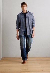 Camisa de botones a rayas azul y blanca, con una camiseta negra debajo, pantalones vaqueros azul deslavado y zapatos marrones, de pie sobre un suelo de madera contra una pared clara.
