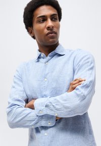 Jaunas vyras, sukryžiavęs rankas, vilkintis šviesiai mėlyną marškinių su sagomis, šiek tiek pažvelgęs į šoną priešais paprastą foną.