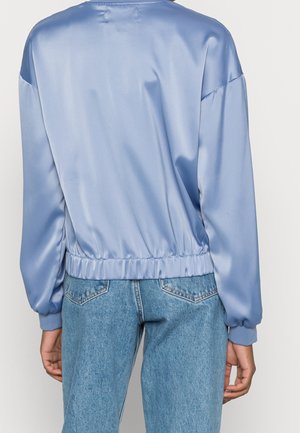 Personne portant un haut bleu clair brillant à manches longues avec une ceinture et des poignets élastiques, associé à un jean en denim bleu taille haute.