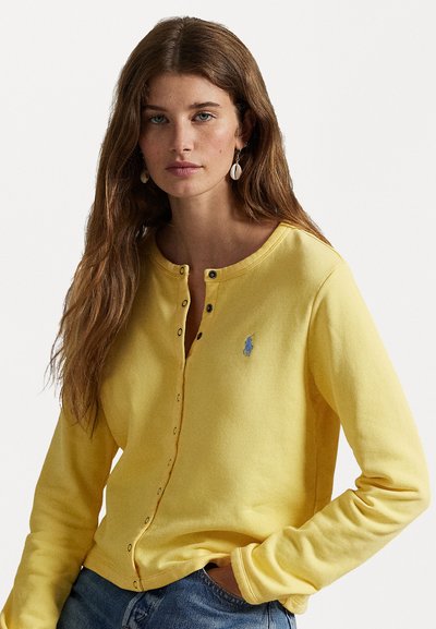 Yellow long-sleeve cardigan made of textured fabric with a button placket and small blue logo. Paired with blue jeans.