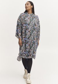 Kobieta w luźnej, kwiecistej sukience do kolan w niebiesko-zielono-brązowych odcieniach, w czarnych legginsach i beżowych butach na sznurówki, stojąca na białym tle.