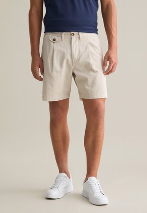 Pantalones cortos beige hechos de una tela texturizada, con pliegues frontales, bolsillos laterales y un pequeño botón decorativo en la cintura.