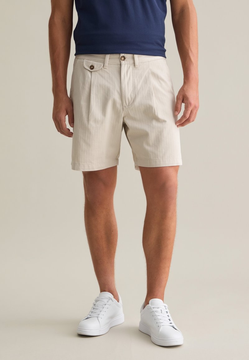 Pantalones cortos beige hechos de una tela texturizada, con pliegues frontales, bolsillos laterales y un pequeño botón decorativo en la cintura.