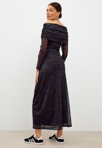 Robe noire à épaules dénudées avec imprimé léopard, corsage froncé, manches longues transparentes et jupe fluide, associée à des baskets noires.