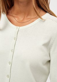 Ljusgrön kofta i mjukt material, med knäppning fram och ribbad halsringning. Utmärker sig för små hålmönster på axlarna.