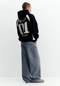 Ung person iført sort hættetrøje med hvid "01" og japansk tekst, kombineret med vidde-bens falmede blå jeans og grå sneakers, stående i profil.
