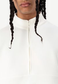 Maglione bianco con zip e colletto a imbuto, caratterizzato da una texture liscia e due ciocche di capelli visibili. Design minimalista con linee pulite.