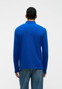 Dlhý rukáv modrá polokošeľa vyrobená z mäkkého materiálu, so golierom a rovnými okrajmi, v kombinácii s svetlomodrými džínsami.