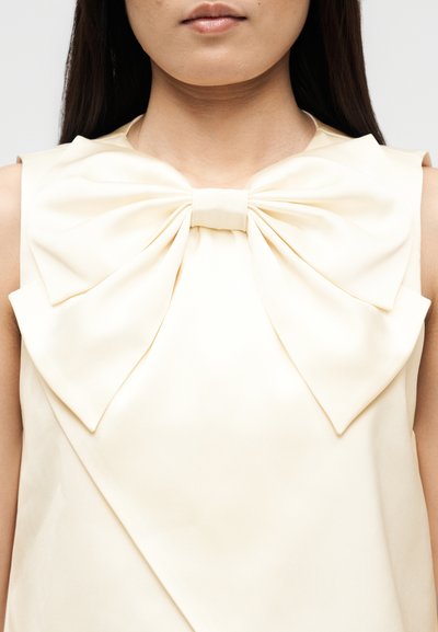 Γυναίκα που φοράει αμάνικο ελεφαντόδοντο τοπ με μεγάλο διακοσμητικό φιόγκο στον λαιμό, με ίσια σκούρα μαλλιά ορατά στα πλάγια.