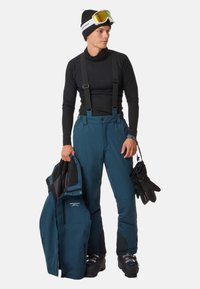 Pantaloni da sci blu con bretelle, una maglia nera a maniche lunghe e una giacca in mano. Gli accessori includono guanti neri e occhiali da sci.