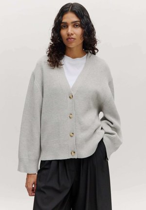 Lichtgrijze cardigan met een V-hals, knoopsluiting aan de voorkant, geribbelde onderkant en verlaagde schouders, gecombineerd met zwarte broek met wijde pijpen.