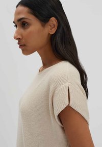 Beiger Strickpullover mit kurzen Ärmeln, der ein strukturiertes Muster und kleine Schlitzdetails an den Ärmelsäumen aufweist, vor einem neutralen Hintergrund präsentiert.