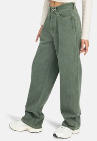 Jean en denim vert surdimensionné avec une taille haute, des coutures visibles et des poches latérales, associé à des chaussures de sport blanches.