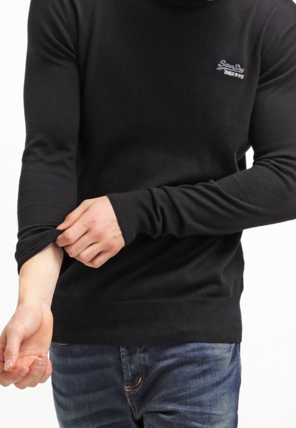 Person in einem schwarzen Langarmpullover mit Logo, der den rechten Ärmel hochkrempelt, kombiniert mit blauen Jeans.