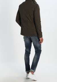 Olivengrøn jakke med høj krave, to brystlommer og en tilpasset pasform, kombineret med blå slim-fit jeans og hvide sneakers.