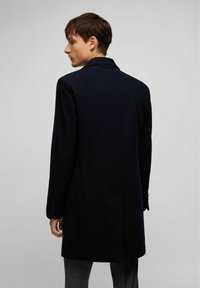 Marineblauer Wollmantel mit einem Reverskragen, langen Ärmeln und einem einzelnen Rückenventil. Mit geknöpften Manschetten und einer insgesamt taillierten Silhouette.