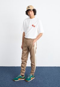 Camiseta blanca con estampado gráfico, pantalones jogger beige con franjas laterales, zapatillas verdes y naranjas, gorra beige. El modelo está de pie frente a un fondo liso.
