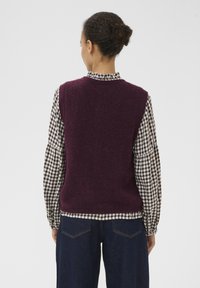 Bordeaux verzwaarde wol zonder mouwen trui met textuur gedragen over een zwart-wit geblokt shirt, gecombineerd met donkerblauwe jeans.