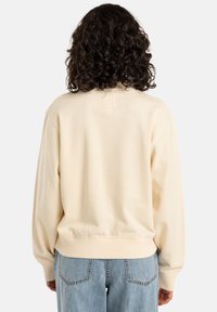 Beige sweatshirt met een zachte textuur, oversized pasvorm, geribde manchetten en een klein stoffen label bij de hals, gecombineerd met lichtblauwe jeans.