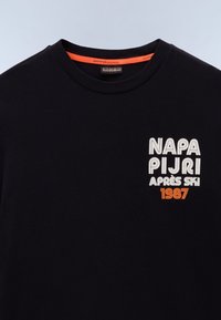 Sort bomulds t-shirt med rund hals. Foran har den hvid og orange broderi: "NAPA PIJRI APRÈS SKI 1987." Glat tekstur.